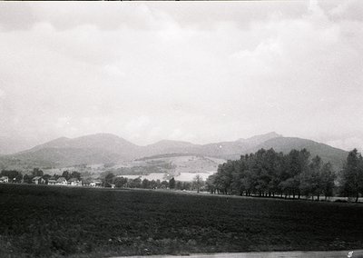 Black-and-white rural landscape featuring rolling hills, dense tree line, and scattered farmhouses. Mid-20th century agricult...