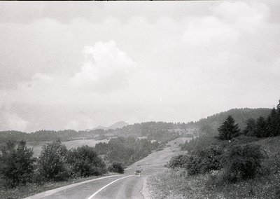 Black-and-white rural road winding through forested hills, likely mid-20th century. Single vintage car on a two-lane asphalt ...