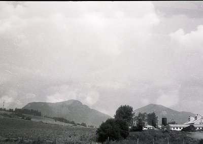 Vintage black-and-white landscape featuring rolling hills, dense forest clusters, and a lone white farmhouse with a cylindric...