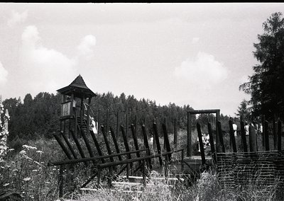 Rustic wooden watchtower with barbed-wire perimeter in dense forest, likely a Cold War-era border outpost. Overgrown vegetati...