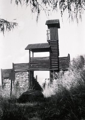 Handcrafted wooden lookout tower with log construction, perched on stone base in forested area. Rustic design features sloped...