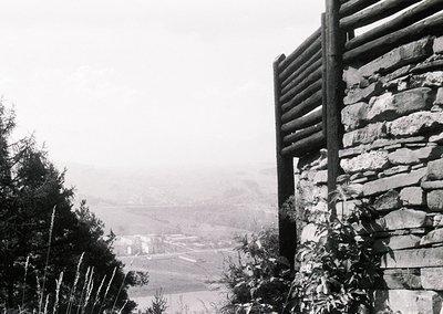 Black-and-white rural scene featuring a stone wall with wooden fence atop a misty valley. Distant farmhouses and fields stret...