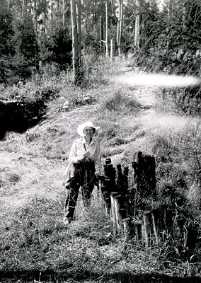 Mid-20th century farmer in rural landscape, seated on wooden fence posts. Dressed in wide-brimmed hat, long-sleeve shirt, and...