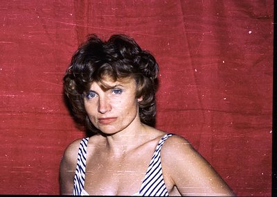 Portrait of a woman in a striped swimsuit against a red backdrop, likely a 1960s–1970s beach or resort setting. Her short, vo...