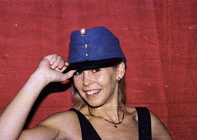Vintage portrait of a woman in a blue military-style cap, sleeveless black top, and layered necklaces. Warm sepia tone sugges...