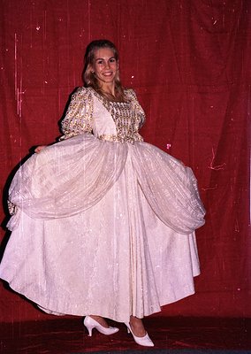 Stage portrait of a performer in an elaborate 1970s-style ballgown, featuring layered tulle skirts and a floral-patterned bod...