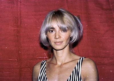 Portrait of a woman with short, layered blonde hair in a 1970s-inspired bob, set against a maroon backdrop. She wears a strip...