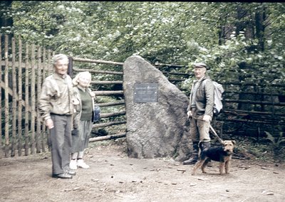 Mid-20th century outdoor scene with three adults and a dog near a wooden fence and large boulder plaque. The man on the right...