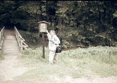 Vintage black-and-white photo of a person posing beside a wooden signpost on a forested trail, likely mid-20th century. The s...