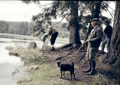 Vintage black-and-white photo of three men and a dog near a forested lakeside, likely mid-20th century. Men in practical outd...