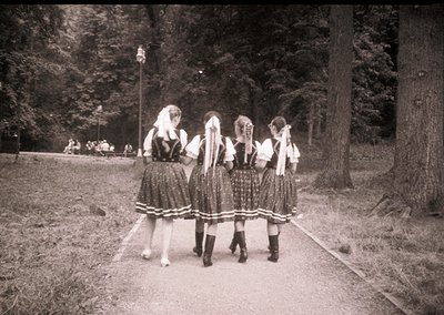 Four women in traditional folk attire—white blouses, dark embroidered skirts, and headscarves—pose outdoors in a wooded park ...