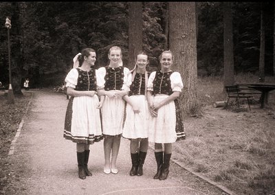 Four women in traditional Alpine folkwear pose on a gravel path, surrounded by dense forest. White blouses with dark embroide...