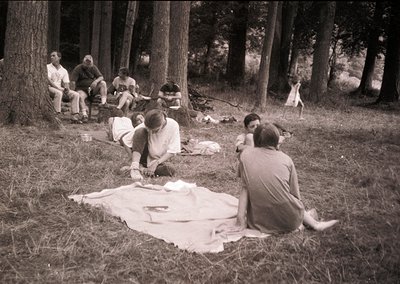 Vintage black-and-white group picnic in a dense forest clearing, mid-20th century. Seven individuals seated/lying on a blanke...