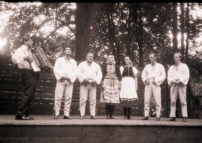 Group of seven performing traditional folk music outdoors, likely Eastern European . Accordionist in white shirt, men in ligh...