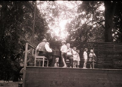 Vintage black-and-white photo of a group of children and an adult on a wooden deck surrounded by dense forest. The adult appe...