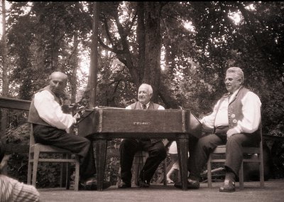 Three men in mid-20th-century attire perform outdoors near dense forest. Left: seated pianist playing a vintage upright piano...