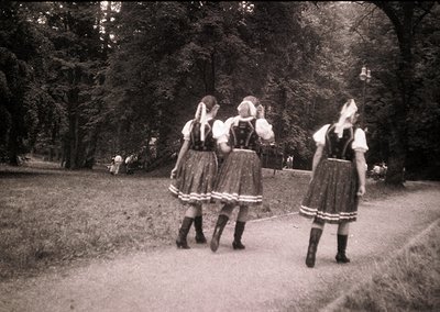 Three women in traditional Bavarian *Dirndl* dresses with white aprons and black knee-high socks walk through a park pathway,...