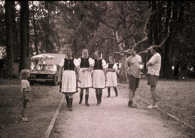 Mid-20th century outdoor gathering in rural setting. Five women in traditional folk attire (blouses, aprons, long skirts) and...