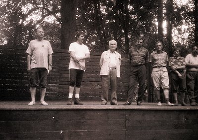 Black-and-white group portrait on a wooden platform, likely 1950s–1960s. Six men in casual attire—short-sleeve shirts, rolled...