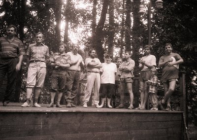 Black-and-white group portrait of eight men posing on a wooden platform in a forested area, likely 1960s–1970s. Casual summer...
