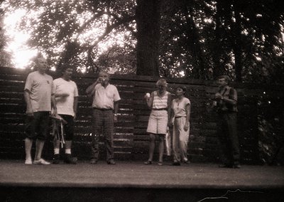 Vintage black-and-white group photo of six adults posing outdoors near a wooden fence. Men wear short-sleeve shirts and trous...