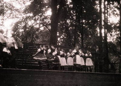 Vintage black-and-white photo of a folk dance ensemble performing outdoors in a wooded area. Women in traditional embroidered...