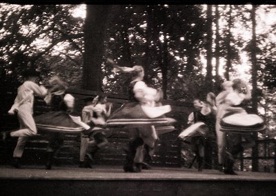Vintage black-and-white photo of a carousel in motion, featuring blurred riders in 1920s-1930s attire. Wooden horses and a ce...