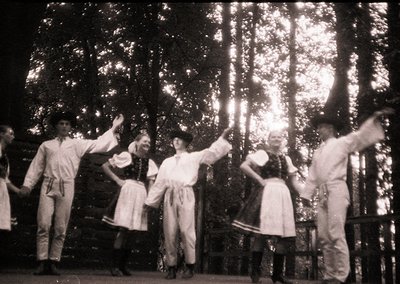 Group performing traditional folk dance in forest setting, likely Eastern European. Men in embroidered vests and women in lon...