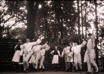 Vintage black-and-white photo of a traditional folk dance in a wooded area, likely Eastern European. Group of 7 dancers in ma...