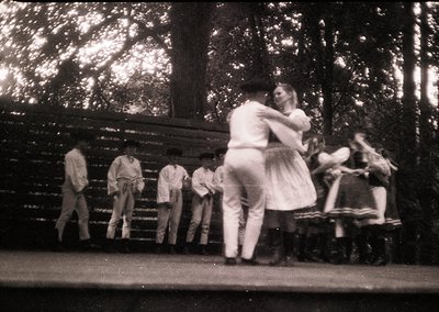 Vintage black-and-white photo of a folk dance performance in a wooded park setting, likely mid-20th century. Couple in tradit...