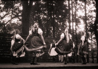 Vintage black-and-white photo of five women in traditional folk costumes performing a circular dance in a wooded area. Elabor...