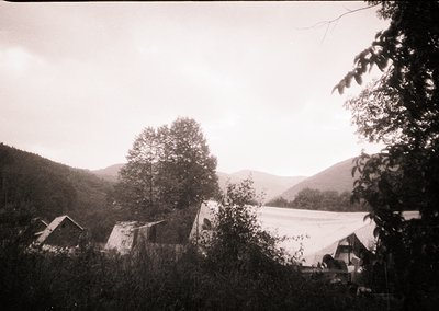 Vintage black-and-white photo of a rural campsite in mountainous terrain, featuring canvas tents and simple wooden structures...