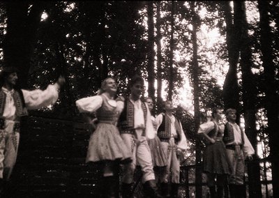 Vintage black-and-white photo of a traditional folk dance performance in a forested setting. Six dancers in period costumes—m...