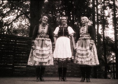 Three women in traditional folk attire pose outdoors, likely Eastern European (1950s–1960s). Intricate embroidered aprons and...