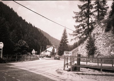 Mid-20th century alpine village roadside with vintage cars, wooden houses, and pine trees. Public phone booth and directional...