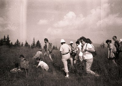 Vintage black-and-white group hiking in grassy alpine terrain, 1960s-70s. Casual attire—wide-brim hats, backpacks, and rolled...