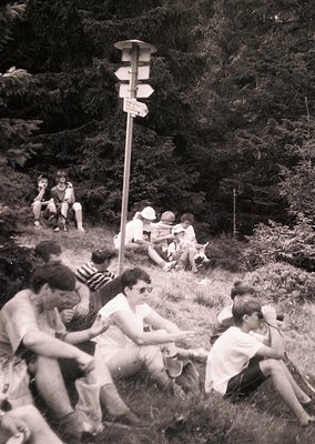 Black-and-white snapshot of a group of young people relaxing in a forested area, likely mid-20th century. Central wooden sign...