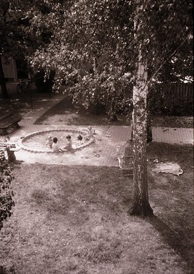 Black-and-white shot of a mid-20th century courtyard poolside scene. Circular concrete pool with four seated figures in swims...