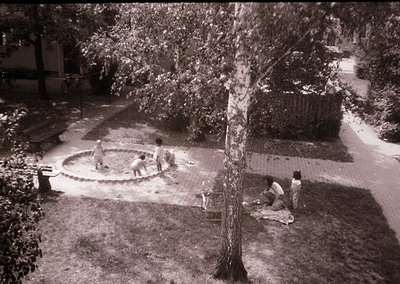 Black-and-white courtyard scene featuring three children playing in a circular concrete water feature. Two children stand at ...