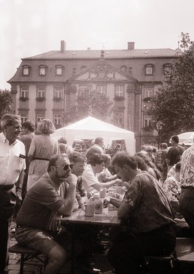 Vintage outdoor gathering at a European café, 1970s. Group of people seated at wooden tables under a white canopy, enjoying d...