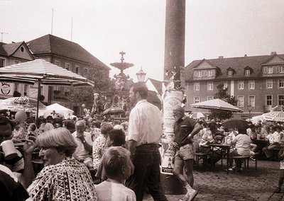 Black-and-white street scene from the 1960s–70s, featuring a lively European town square with mid-century architecture. Crowd...