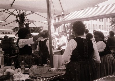 Vintage outdoor café scene with women in early 20th-century attire, likely 1920s–1930s. Striped awnings, vintage signage ("Pe...