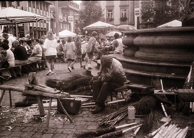 Street vendor sharpening tools beside cobblestone plaza. Mid-20th century European café scene with umbrellas and outdoor seat...