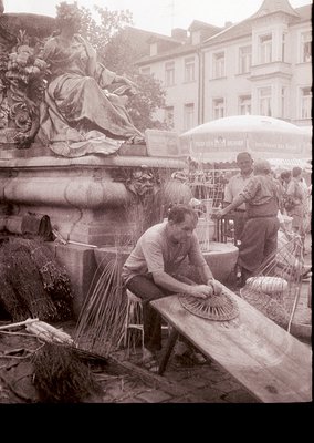 Vintage black-and-white street scene featuring two artisans crafting traditional straw hats on a wooden bench beside a classi...