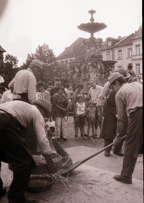 Vintage street scene featuring a group of men clearing debris around an ornate, multi-tiered fountain in a European town squa...