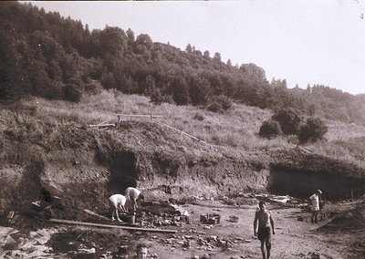 Black-and-white photo of early 20th-century tunnel construction in a forested mountainous region. Workers in period attire us...