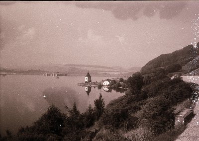 Vintage sepia-toned lakeside village with a prominent church tower on a small island. Surrounding greenery and rolling hills ...