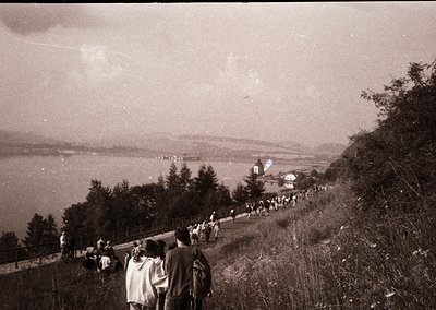 Black-and-white shot of a coastal hillside path crowded with mid-20th-century attire (1950s–60s). Group of hikers descends to...