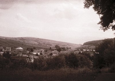 Vintage sepia-toned rural village nestled in rolling hills, likely Eastern Europe. Clustered brick homes with pitched roofs a...