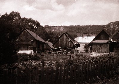 Vintage sepia-toned rural farmstead with timber-framed houses, likely Eastern European . Wooden fences, stacked firewood, and...
